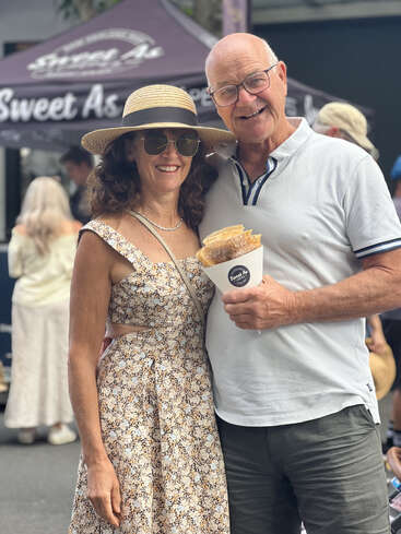 Una pareja sonriente posa en un evento al aire libre. La mujer lleva gafas de sol, un sombrero para el sol y un vestido de flores. El hombre sostiene caramelos, ambos parecen felices.