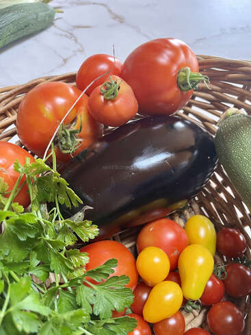 Una cesta de mimbre llena de verduras frescas: tomates rojos y amarillos, una berenjena, calabacines, perejil y tomates cherry, sobre una encimera de mármol.