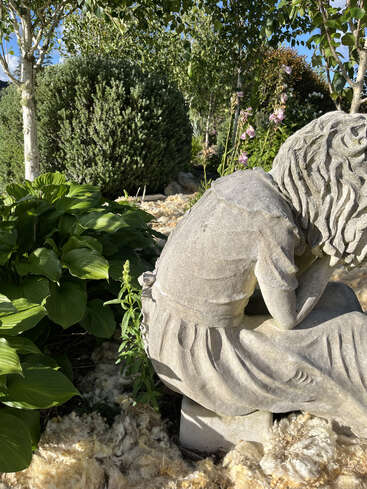 Una estatua de piedra de una niña está sentada con la cabeza sobre las rodillas en un jardín, rodeada de exuberante vegetación, arbustos y la luz del sol filtrándose entre los árboles.