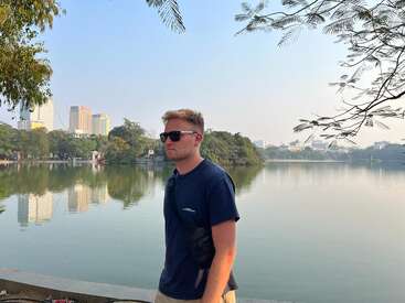 Un hombre con gafas de sol y camiseta azul marino está de pie junto a un tranquilo lago. Los edificios de la ciudad y los frondosos árboles se reflejan bellamente en el agua.