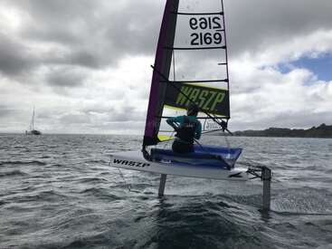 Una persona navega con un hidrodeslizador WASZP por encima del agua en un día nublado, con el velero lejano y la costa a la vista, demostrando velocidad y equilibrio.