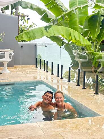 A smiling couple enjoys a small pool, surrounded by elegant tiles and lush banana leaves. Glass fencing offers a scenic view of water and greenery.