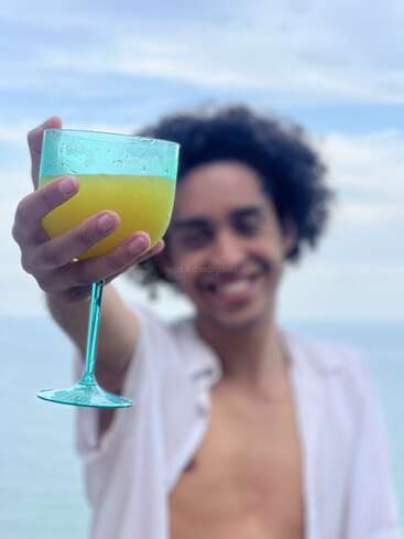 A smiling person holds a blue stemmed glass filled with a yellow drink towards the camera, standing outdoors with the sky and water in the background.