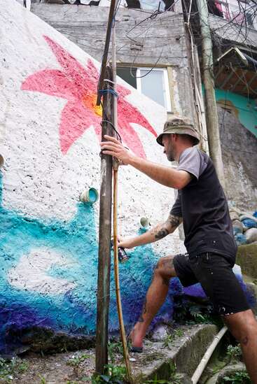 A man wearing a bucket hat and black clothes paints a colorful mural of a flower on a textured outdoor wall, using spray paint, in an urban setting.