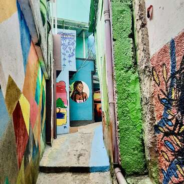 A narrow alleyway features vibrant murals on every wall, including geometric patterns, colorful textures, and a painted portrait of a woman at the end. Urban art.