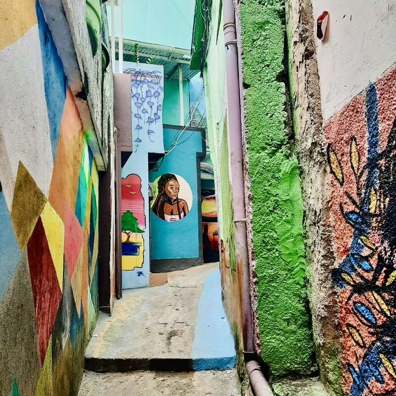 A narrow alleyway features vibrant murals on every wall, including geometric patterns, colorful textures, and a painted portrait of a woman at the end. Urban art.