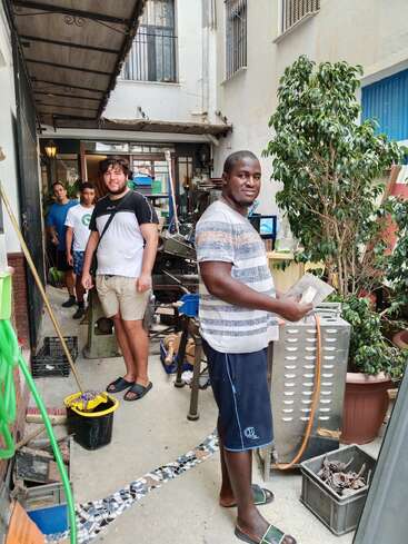 Quatre hommes se tiennent dans un atelier ou une cour encombrée, entourés d'outils, de plantes et de matériel. Ils semblent travailler ou préparer un projet ensemble.