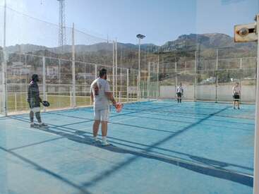 Quatre personnes jouent au padel sur un terrain bleu entouré d'une clôture transparente, avec des montagnes et des bâtiments visibles sur le fond clair et ensoleillé.