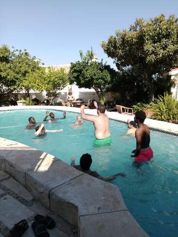Un groupe de personnes joue avec une balle colorée dans une piscine, entourée d'arbres, de meubles d'extérieur et d'un soleil radieux, profitant d'une journée d'été.