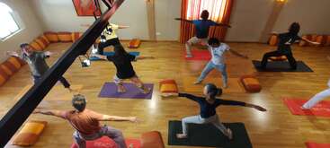 Un groupe de personnes pratique le yoga dans un studio bien éclairé, avec un sol en bois, des tapis colorés et des coussins. Tout le monde tient la posture du guerrier, en se concentrant sur l'équilibre.