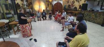 Un groupe de personnes est assis en cercle dans une pièce rustique, écoutant attentivement une femme debout qui parle, créant ainsi une atmosphère concentrée et interactive.