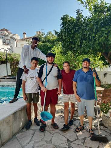 Cinq hommes se tiennent près d'une piscine en plein air, avec des outils de nettoyage à la main. Ils sont souriants, profitant d'un temps ensoleillé, entourés d'arbres, de verdure et de bâtiments méditerranéens blancs.