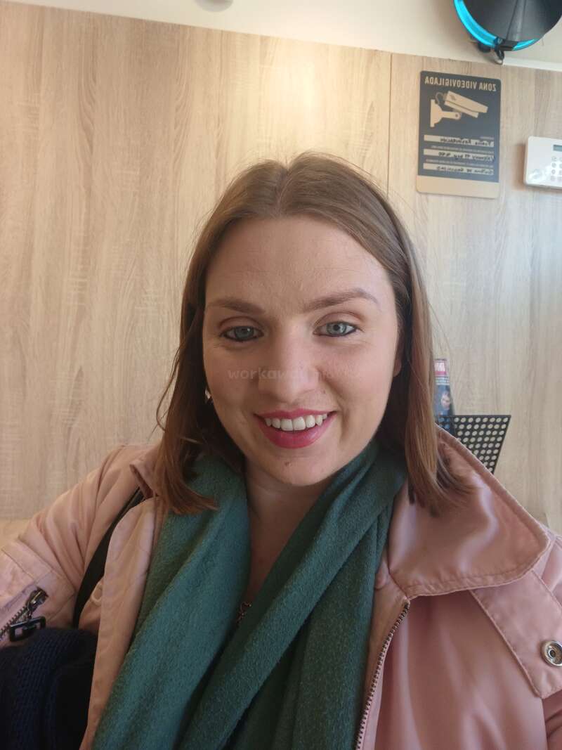 A woman with light brown hair smiles at the camera, wearing a pink coat and green scarf. Wooden wall and security camera sign visible in the background.