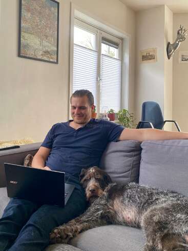 Ein Mann sitzt bequem auf einer grauen Couch mit einem Laptop und lächelt. Sein Hund liegt entspannt neben ihm. In dem gemütlichen Raum gibt es Pflanzen, Kunst und Sonnenlicht.