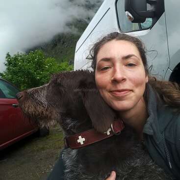 Eine Frau lächelt und umarmt einen braunen, struppigen Hund. Sie sind im Freien in der Nähe von Fahrzeugen, mit Bäumen und nebligen Bergen im Hintergrund. Ein gemütlicher, natürlicher Moment.