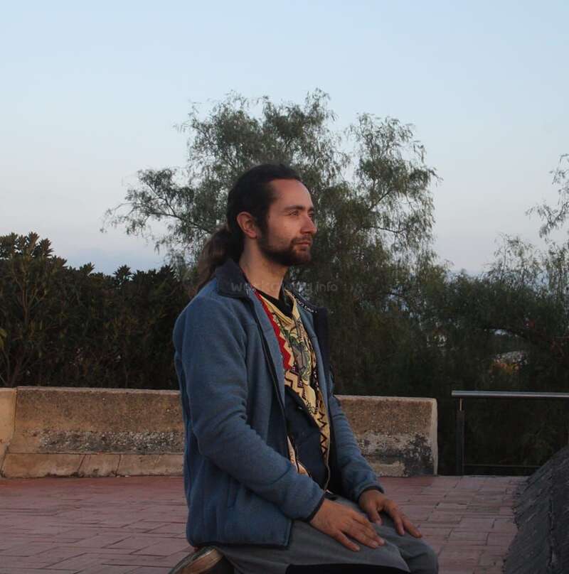 Un hombre con el pelo largo está tranquilamente sentado de rodillas al aire libre, vestido con una chaqueta azul. Al fondo, árboles y un muro de piedra bajo un cielo despejado.