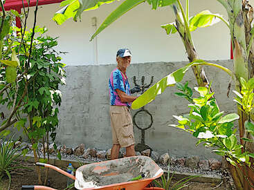 Ein Mann in einem bunten Hemd und einer Mütze steht in einem üppigen Garten und lächelt, während er eine Figur auf eine Zementwand hinter sich malt oder zeichnet.