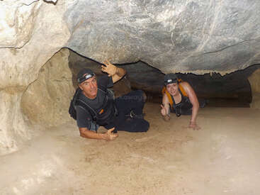 Zwei Menschen krabbeln mit Helm und Rucksack durch einen engen Höhlengang. Die felsige Decke ist niedrig, sodass sie sich auf Händen und Knien fortbewegen müssen.