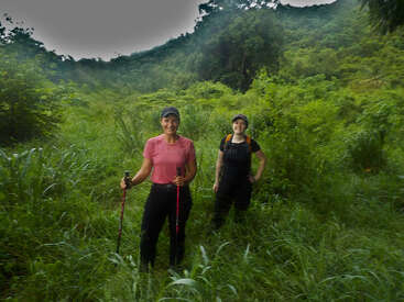 Zwei Frauen wandern durch eine üppige, grüne Vegetation in einem Waldgebiet. Beide lächeln, tragen Hüte und sind in Outdoor-Kleidung gekleidet und genießen gemeinsam die Natur.