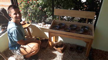 A person is smiling while kneeling indoors near a wooden sensory table with metal bowls. Straw is spread on the floor. Greenery is visible outside.