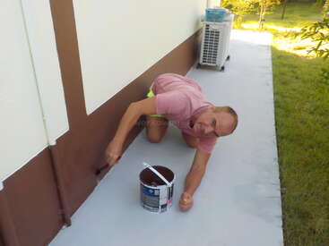 A man in a pink shirt and yellow shorts kneels on the ground, painting the base of a building brown, next to a paint can, outside.