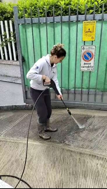Una mujer con botas y sudadera con capucha está utilizando una hidrolimpiadora para limpiar la entrada de una casa, situada frente a una verja metálica, donde se ven señales de advertencia y de prohibición de aparcar.
