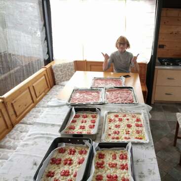 Una mujer está sentada en una mesa de madera en una acogedora cocina, posando orgullosa junto a varias bandejas de focaccia recién hecha y pizzas listas para hornear.