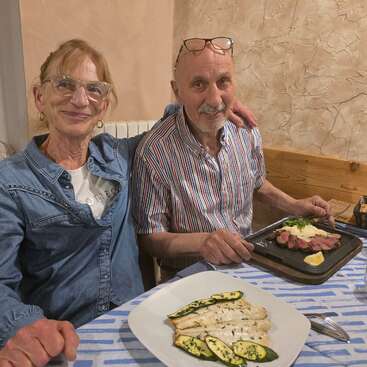 Una pareja de ancianos está sentada en la mesa de un restaurante, sonriendo con cariño. Tienen ante sí unos deliciosos platos de pescado a la plancha con calabacín y carne en tiras con verduras.