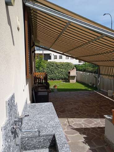 Un patio con suelo de piedra, un fregadero exterior de granito y una marquesina a rayas que da sombra. Se puede ver un césped verde, una barbacoa, sillas y una valla que lo rodea.
