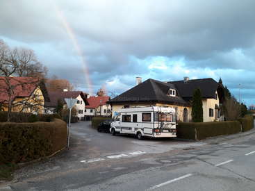 The image depicts a serene residential street with a white camper van parked on the side of the road, surrounded by houses and a vibrant rainbow in the sky.