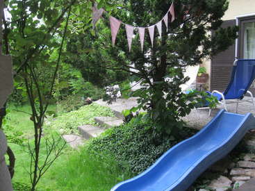 The image depicts a serene backyard scene featuring a blue slide, a tree, and a patio area, with lush greenery and a festive bunting.