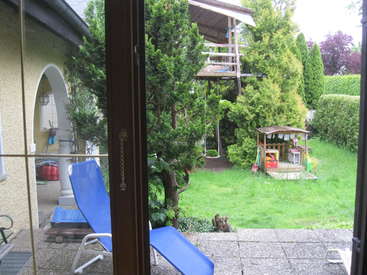 The image depicts a serene backyard scene, featuring a house, a blue lounge chair, a playhouse, and lush greenery, captured through a glass door.