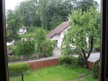 The image depicts a serene residential area, featuring a white house with a brown roof, surrounded by lush greenery and a fence, viewed from a window.