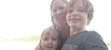 A woman and two children, likely a family, pose closely together for a selfie outdoors. The background appears bright, possibly a sunny day near water or mountains.