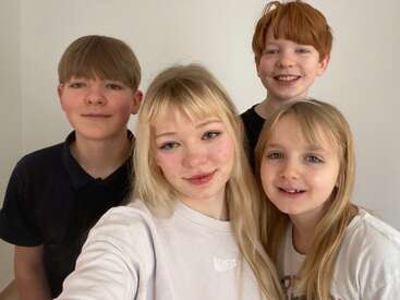 Four children pose together in a brightly lit room, smiling warmly at the camera. They appear happy and comfortable, suggesting a close relationship or sibling bond.