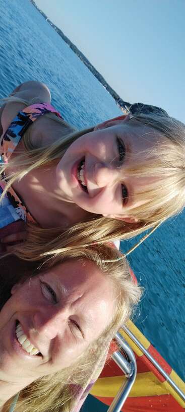 A woman and young girl smile together on a sunny day, enjoying time by the water, possibly on a boat, with clear blue sky and sea.