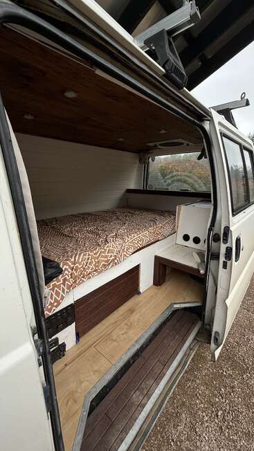 This image shows the inside of a converted van with a cozy bed, wooden ceiling, and floor, white walls, and a small built-in storage area.