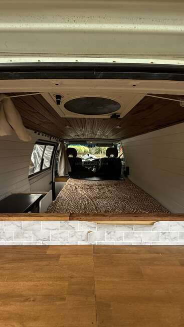 Essa imagem mostra o interior de uma van reformada. Tem um teto de madeira, uma cama estampada, uma parte com azulejos, uma pequena cozinha e os bancos da frente visíveis. Um ambiente bem aconchegante.