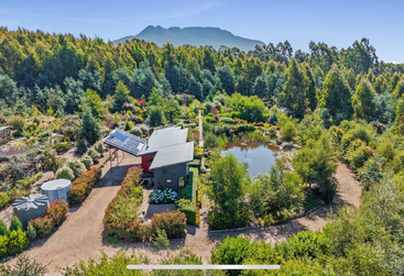 Uma propriedade rural super charmosa tem uma casa moderna com painéis solares, cercada por jardins lindos, um lago, caminhos circulares, tanques de água e montanhas florestadas ao fundo.