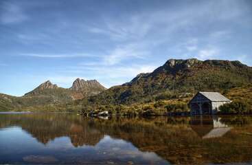 Um lago tranquilo reflete montanhas escarpadas sob um céu azul, com um pitoresco barracão de madeira à beira da margem, cercado por uma vegetação exuberante e uma beleza natural tranquila.