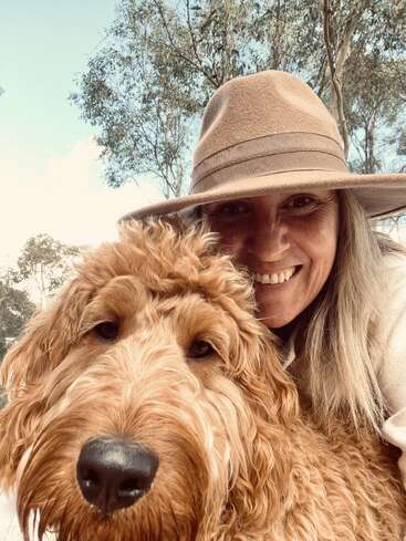 Uma mulher feliz, usando um chapéu de abas largas, sorri ao lado de seu cachorro fofo e dourado. Eles parecem estar ao ar livre, curtindo um momento caloroso e alegre juntos.