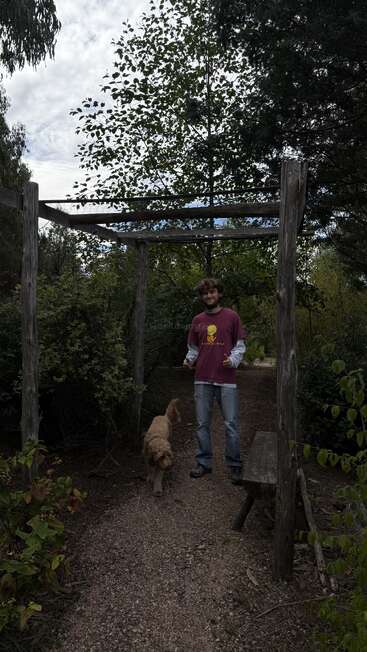 Um rapaz está debaixo de um arco rústico de madeira, num caminho de jardim bem verdejante, vestindo jeans e uma camisa marrom-avermelhada, acompanhado por um cachorrinho de cabelos cacheados.