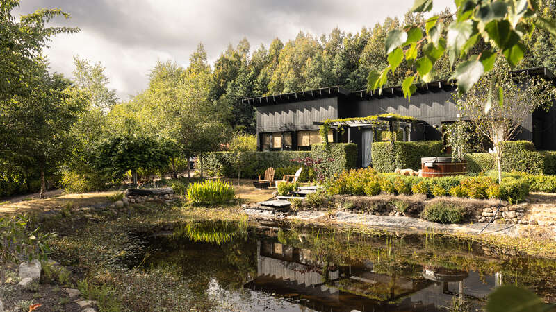 Uma casa preta moderna fica cercada por uma vegetação exuberante e árvores, com um lago tranquilo em primeiro plano refletindo o jardim e a arquitetura pacíficos.