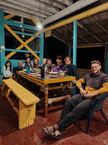 Neuf personnes sont assises autour d'une table en bois rustique sous un patio couvert, partageant un repas et des sourires, profitant d'une soirée confortable, détendue et communautaire.