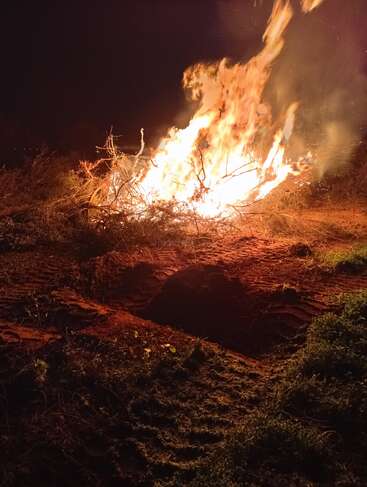 A large, bright bonfire burns intensely at night, consuming dry branches and twigs. The flames illuminate the surrounding earth and cast dramatic shadows nearby.