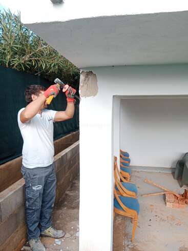 A man wearing gloves and work clothes uses a hammer and chisel to repair or demolish a damaged corner of a concrete structure near some chairs.
