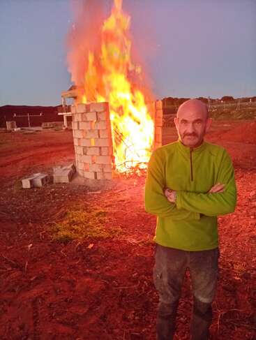 Ein Mann in grüner Jacke steht mit verschränkten Armen vor einem großen, lodernden Feuer, das von einem gemauerten Gebäude auf rötlichem Boden im Freien eingeschlossen wird.