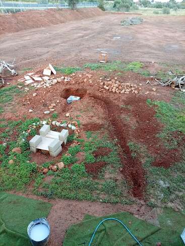 A freshly dug hole with scattered stones, some construction blocks forming a small structure, a bucket with liquid, and a hose on earthy and grassy ground.