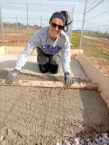 A woman wearing sunglasses and gloves kneels on the ground, smoothing freshly poured concrete inside a fenced area, smiling and enjoying her outdoor construction work.