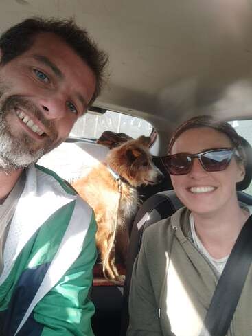 A man and woman are smiling in a car selfie, with a brown dog sitting in the back seat. Sunlight brightens their happy, relaxed faces.